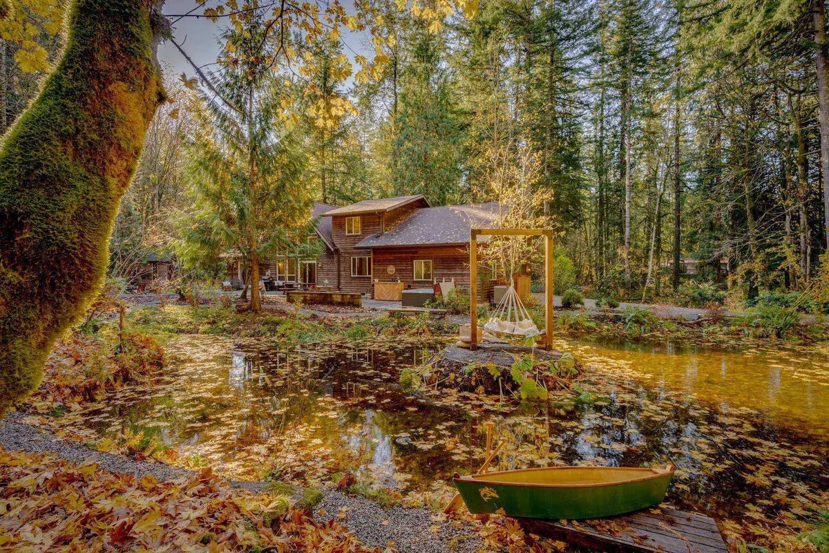A tranquil pond reflecting the green hues of the surrounding forest, with a wooden cabin nearby and a green canoe invitingly positioned on the shore. A swing set adds a playful touch while the overall ambiance feels peaceful and secluded.