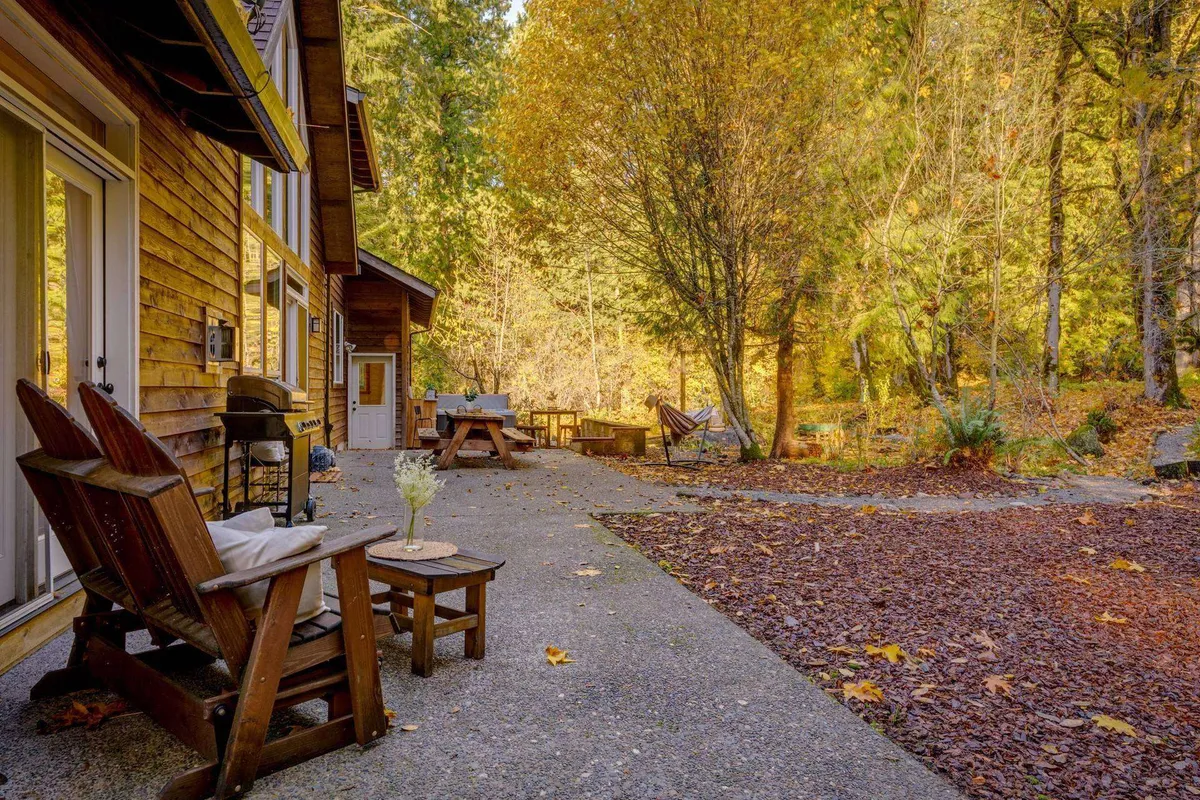 An inviting outdoor area with Adirondack chairs circling a wooden table, a gently swaying hammock strung between the trees, and a pathway leading into the woods. The setting is serene, perfect for socializing, reading, or watching the sun set in the company of friends and family.
