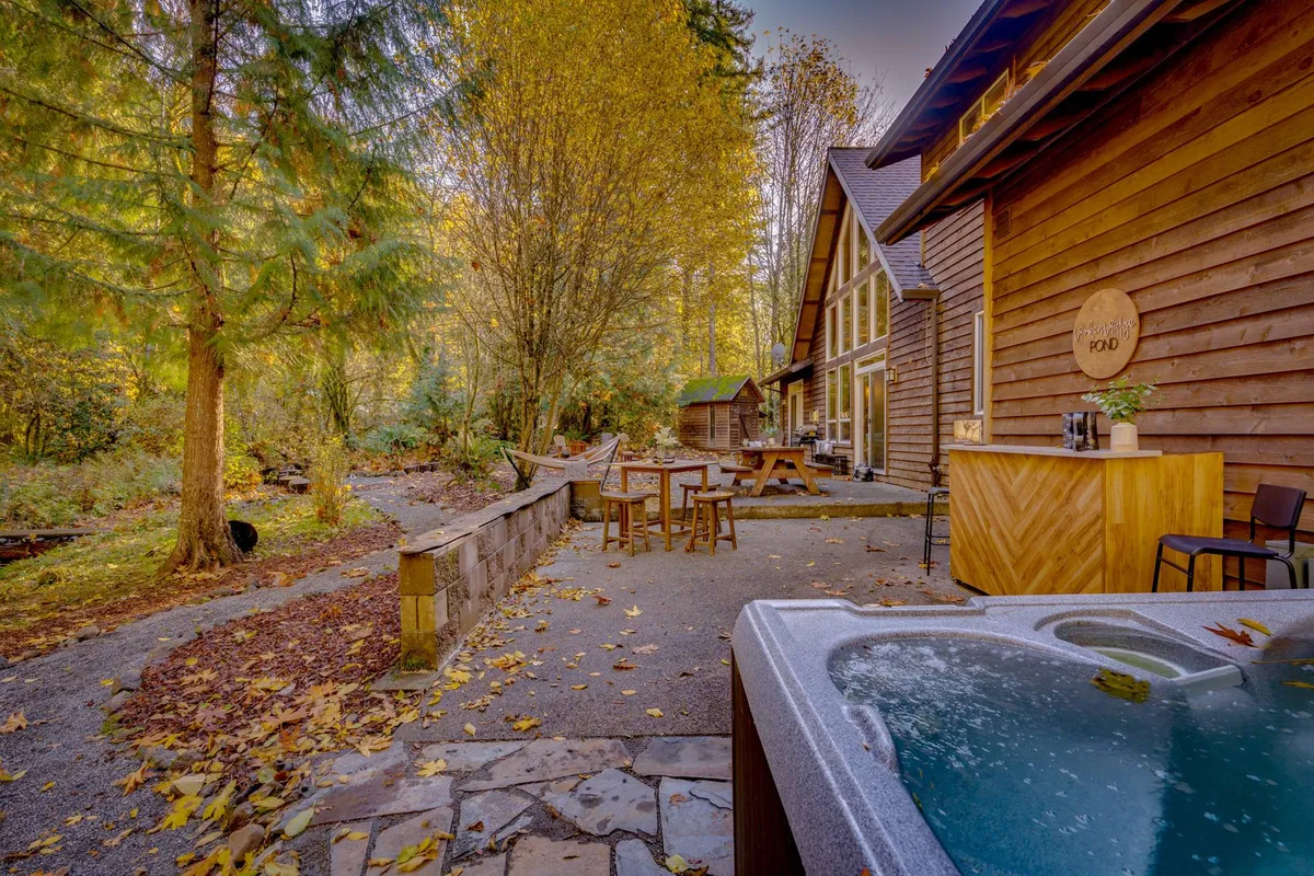 A tranquil exterior scene featuring a bubbling hot tub on a wooden patio, surrounded by lush trees. A rustic stone wall and a wooden patio table add to the relaxing ambiance, creating an ideal space for stargazing and group gatherings after a day outdoors.