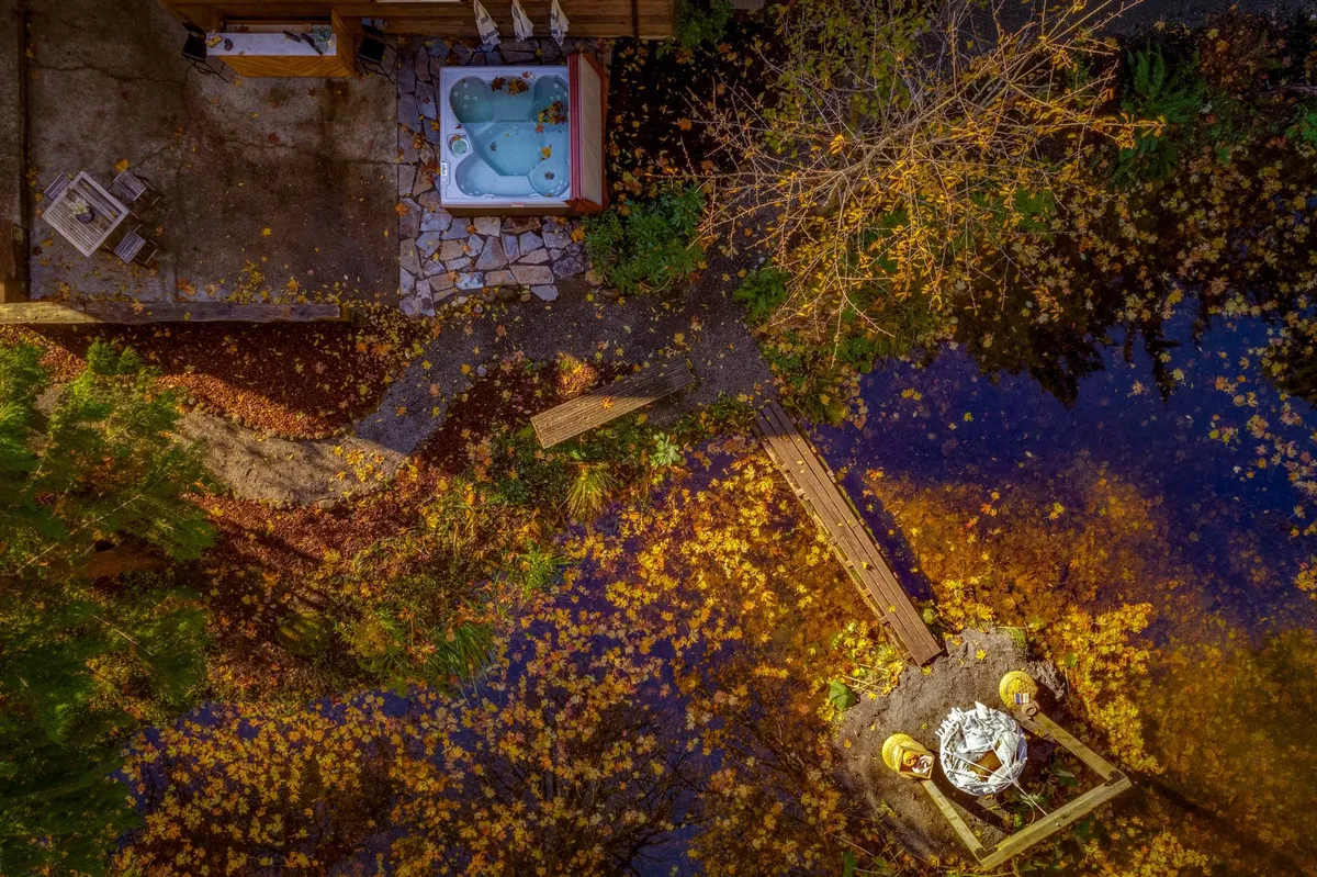 A spacious wooden deck bordered by lush garden areas and golden yellow leaves, featuring a bubbling hot tub, outdoor seating arrangements, and a serene backdrop of nature. The inviting layout showcases both dining and relaxation zones, perfect for social gatherings and quiet moments alike.
