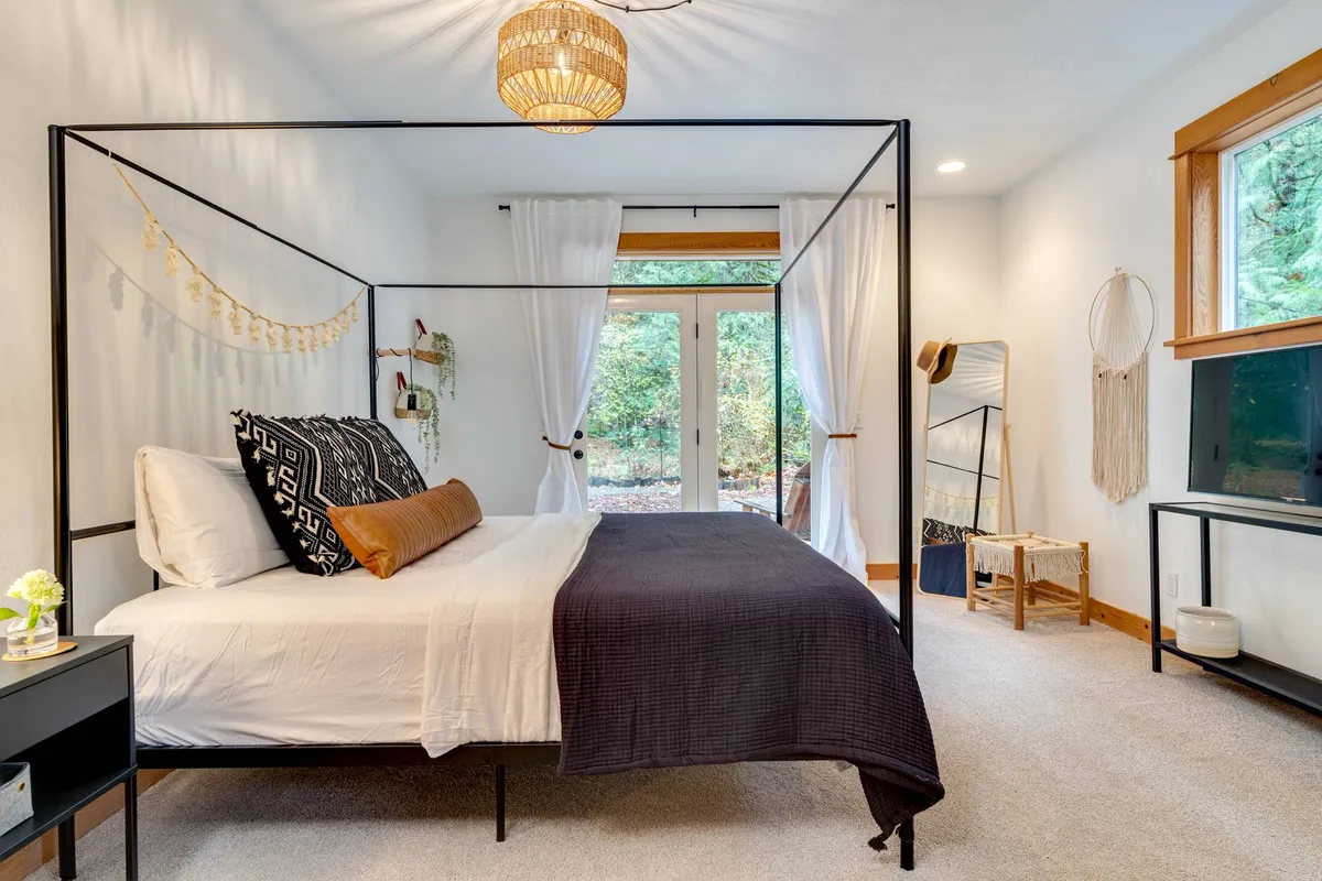 A tranquil bedroom with a canopy bed, textured throws, and natural light streaming in through glass doors. Decorative wall hangings add warmth, and the peaceful atmosphere is ideal for rest or romantic escapes. The view suggests easy access to a private patio or deck area, aligning with the Cedar suite's description.