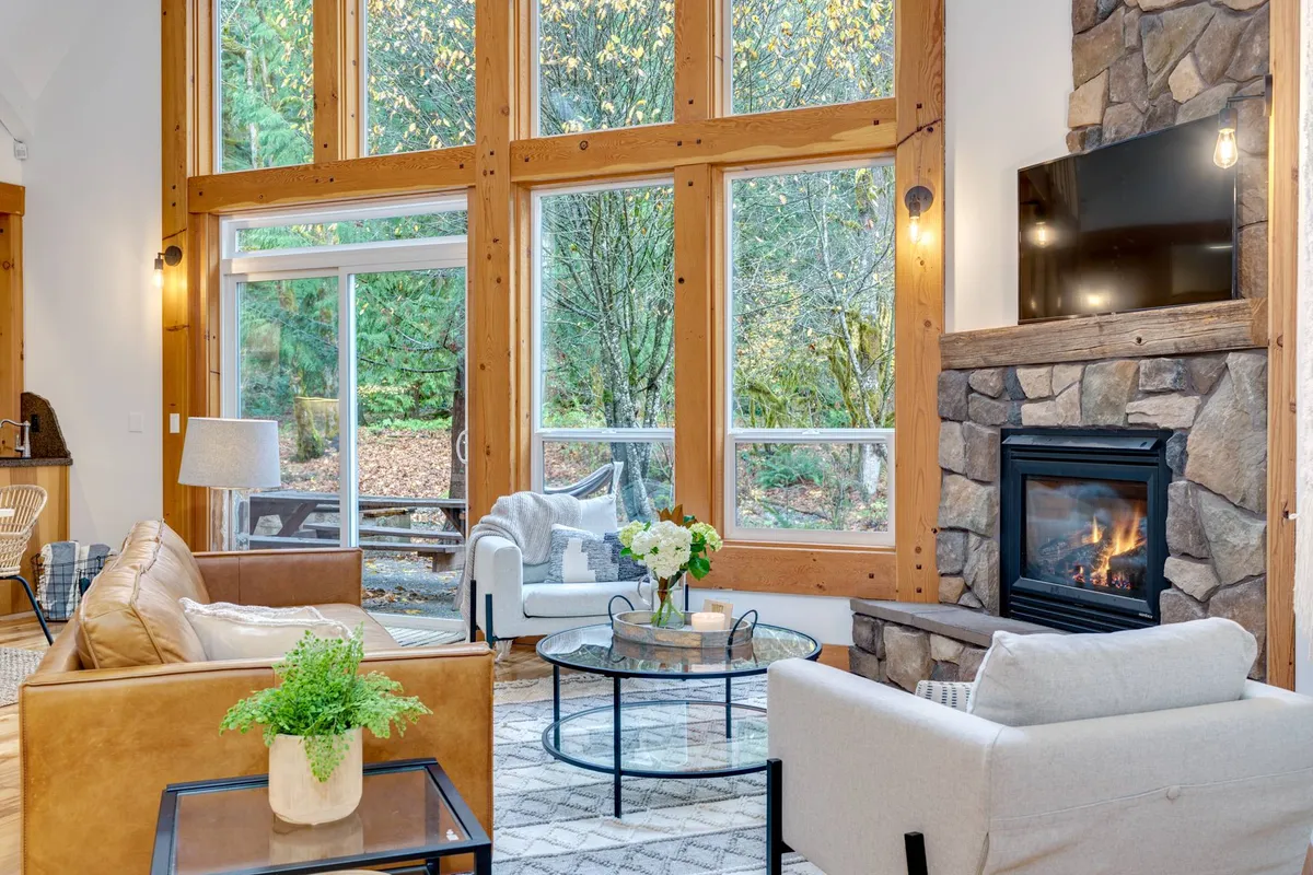 A spacious and welcoming living area featuring large windows that flood the room with natural light, offering glimpses of the surrounding landscape. The stone fireplace and comfortable seating invite relaxation after a day of adventure, while a coffee table with fresh flowers adds a homey touch.