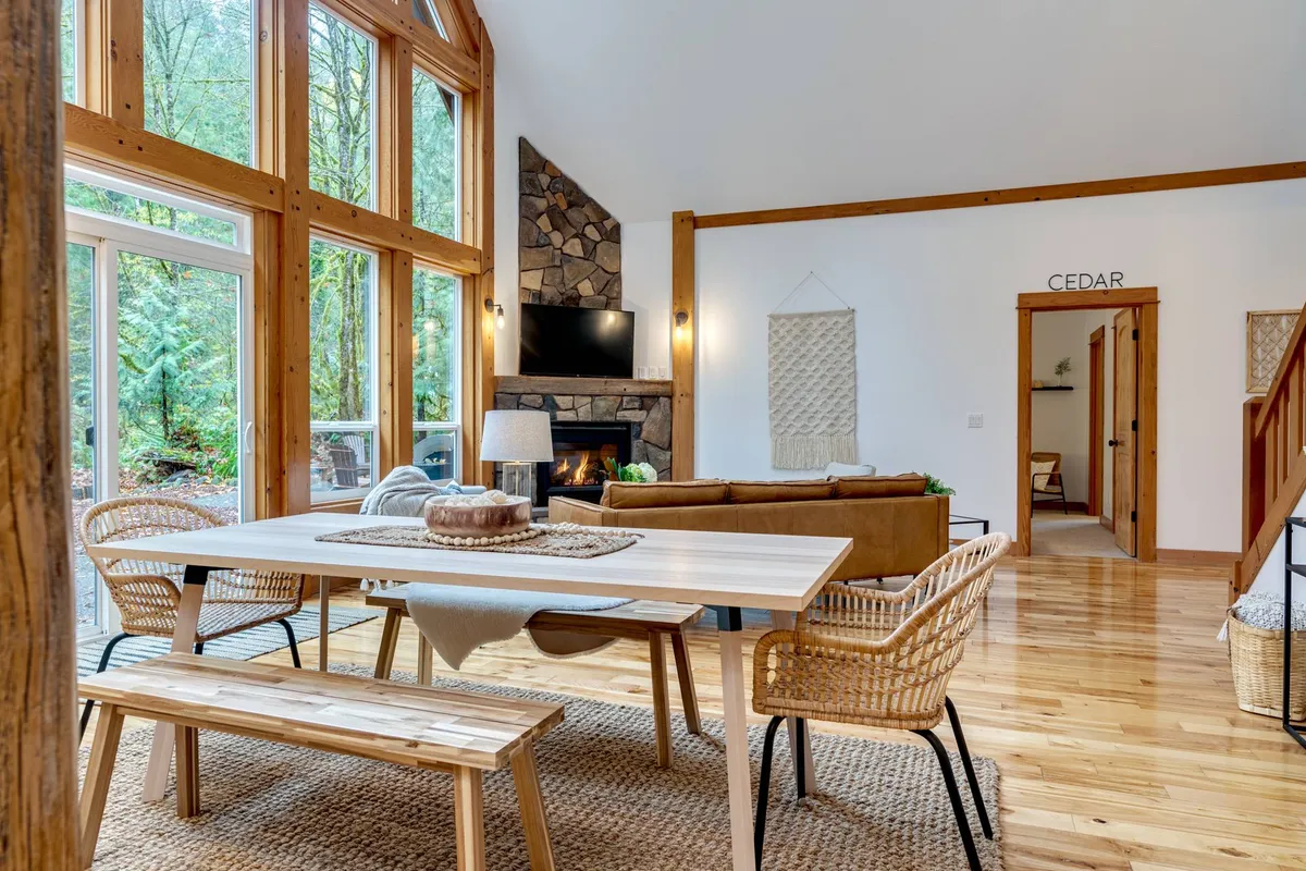 A cozy dining and living area bathed in natural light from the expansive windows, which frame views of the forested surroundings. The stone fireplace creates a warm ambiance, perfect for sharing stories after a day exploring the mountain.