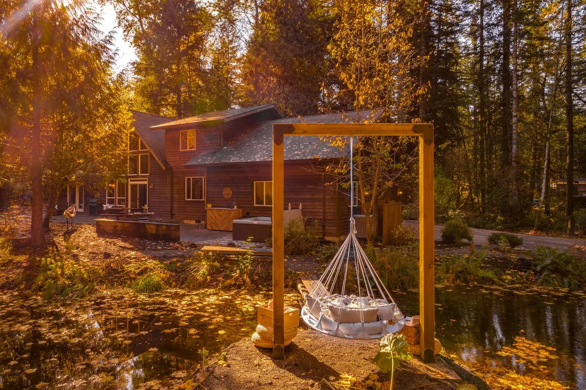 The photo highlights the house overlooking a tranquil pond, with distinctive hanging seating and vibrant fall foliage. The unique seating area by the water and colorful surroundings create a welcoming environment for both relaxation and adventure, perfectly suited for family gatherings and child-friendly exploration.