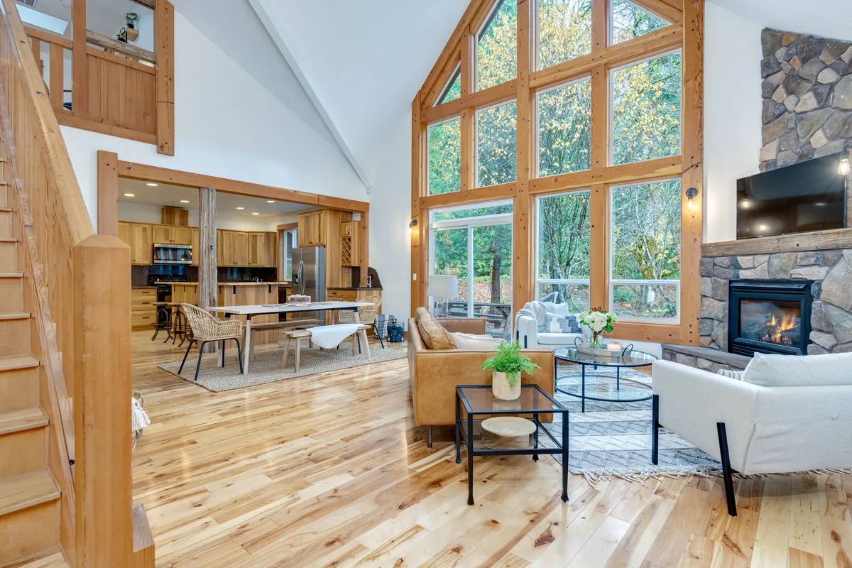 This image captures a spacious and airy open-concept living area featuring large windows that flood the space with natural light, a prominent fireplace, a wooden staircase, and an adjacent kitchen. The arrangement of furniture and warm tones accentuate the inviting and communal feel.