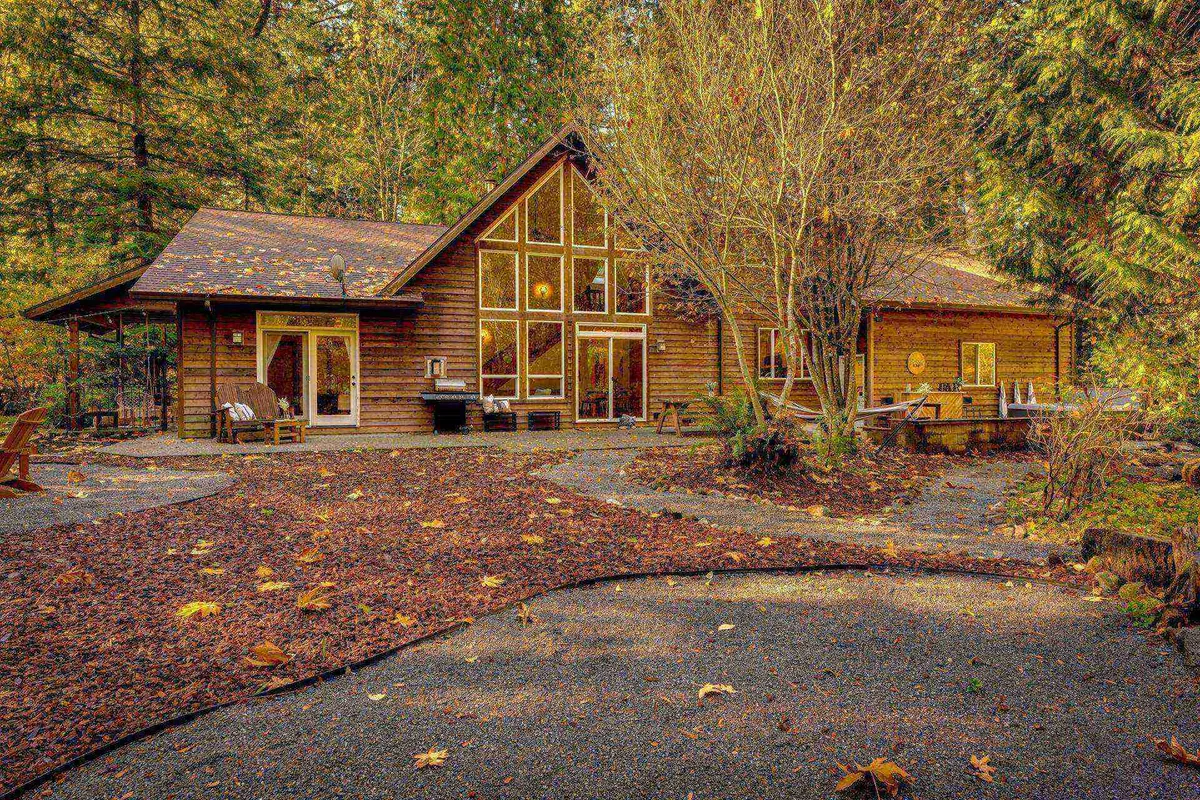 This image features the exterior of the home, showcasing its large windows and wooden facade nestled among tall fir trees. A winding pathway leads toward the house, while the lush greenery and surrounding trees evoke a sense of privacy and peacefulness.