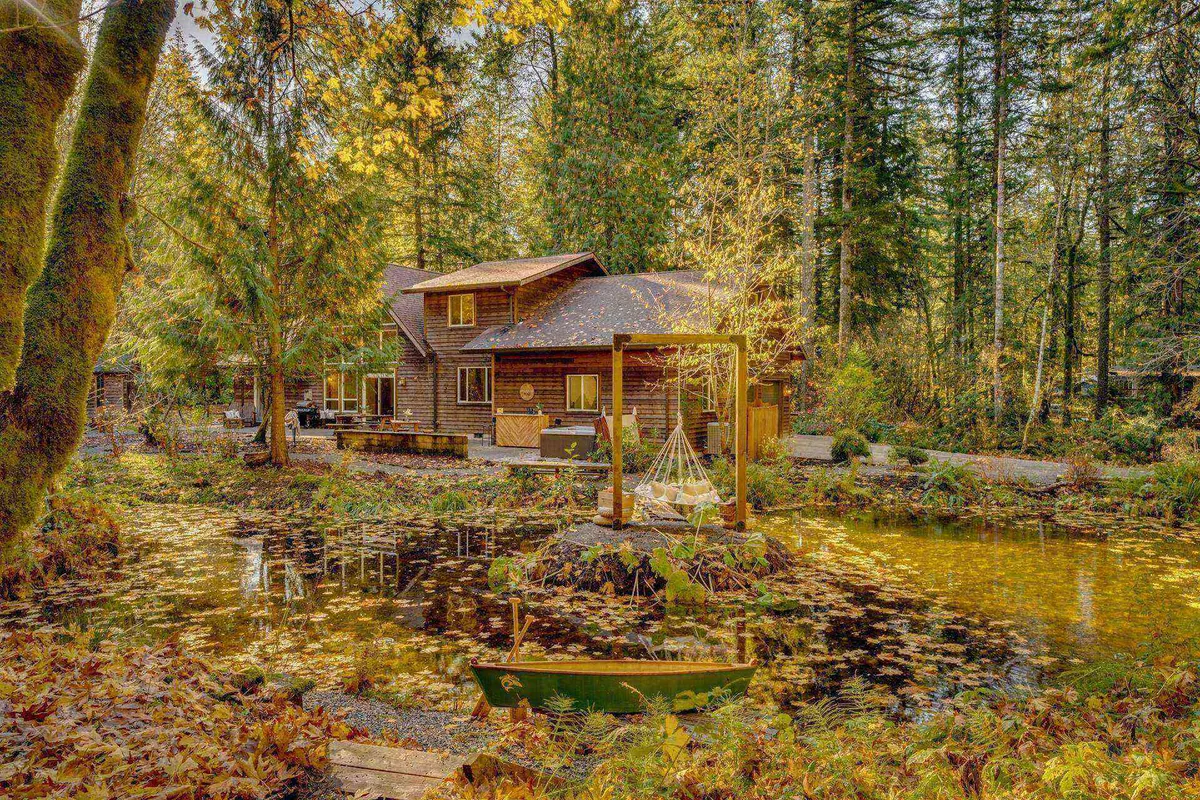 This exterior image features the wooden house nestled among towering trees, a hammock strung between trunks, a serene pond, and lush natural surroundings. The scene captures multiple outdoor amenities, including the pond suitable for canoeing or fishing and communal relaxation spots, perfectly evoking the property's private, adventure-filled landscape.