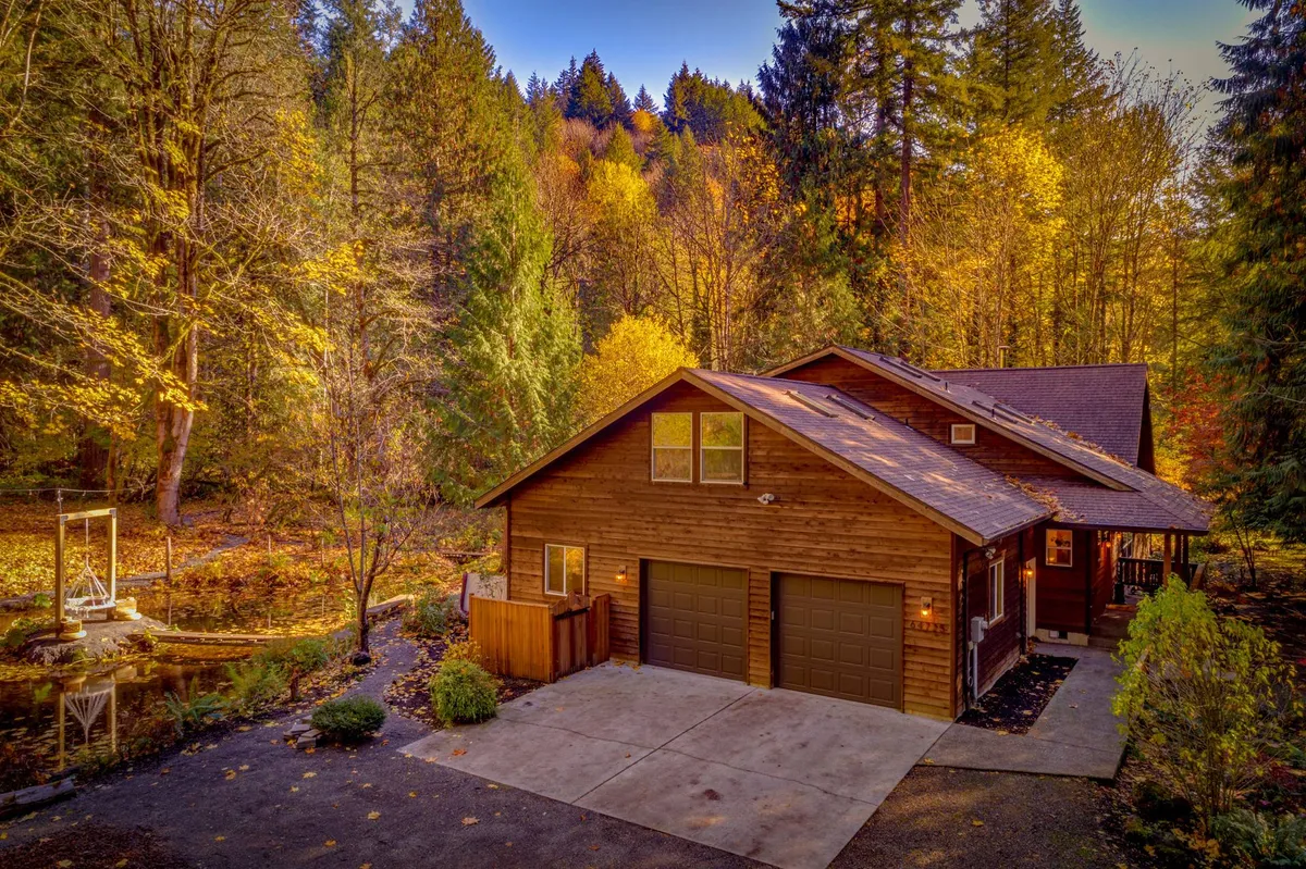 A beautiful exterior view of a wooden house framed by vibrant autumn foliage, a tranquil pond, and a spacious driveway. The warm glow of the setting sun bathes the scene, highlighting the peaceful and private natural surroundings.