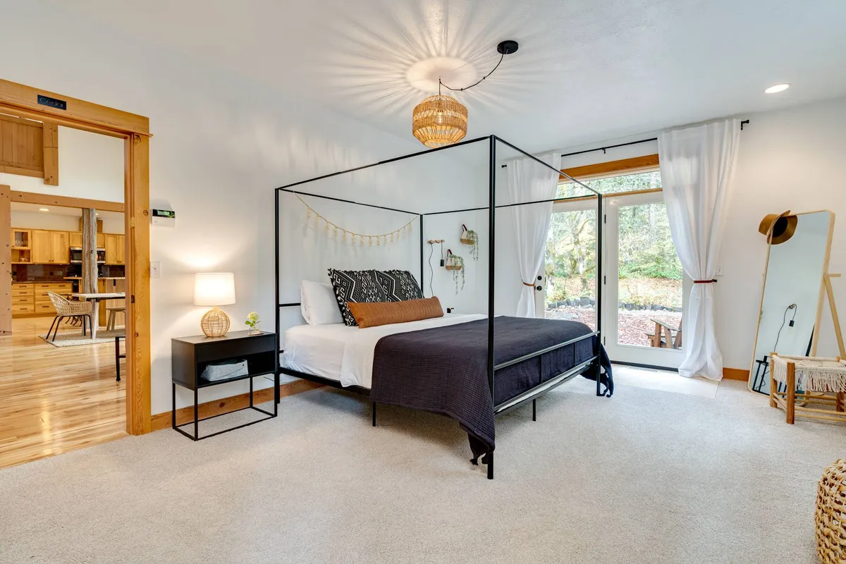A bright, serene bedroom with a four-poster bed and large windows showcasing views of nature. The decor is clean and inviting, with a comfortable nightstand and a decorative light fixture, suggesting a restful ambiance for guests.