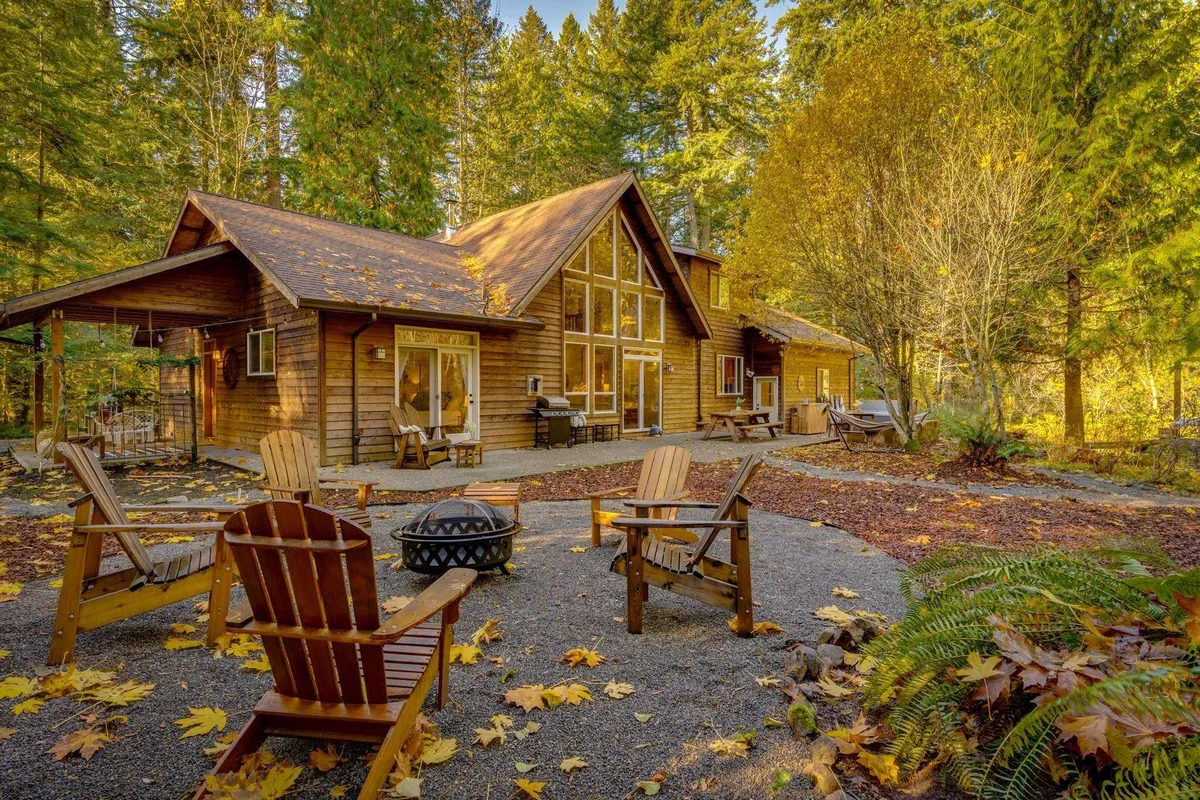 The hero image showcases a beautifully remodeled wooden cabin nestled among towering trees, with a welcoming fire pit surrounded by Adirondack chairs in the spacious yard. The tranquil forest setting, inviting outdoor seating, and soft sunset light evoke a cozy, secluded retreat ideal for gatherings and relaxation.